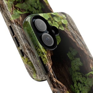 May include: A close-up of a phone case with a forest scene. The case features a dark brown tree trunk with green moss and ferns growing on it. The phone's camera lens is visible in the upper right corner of the image.