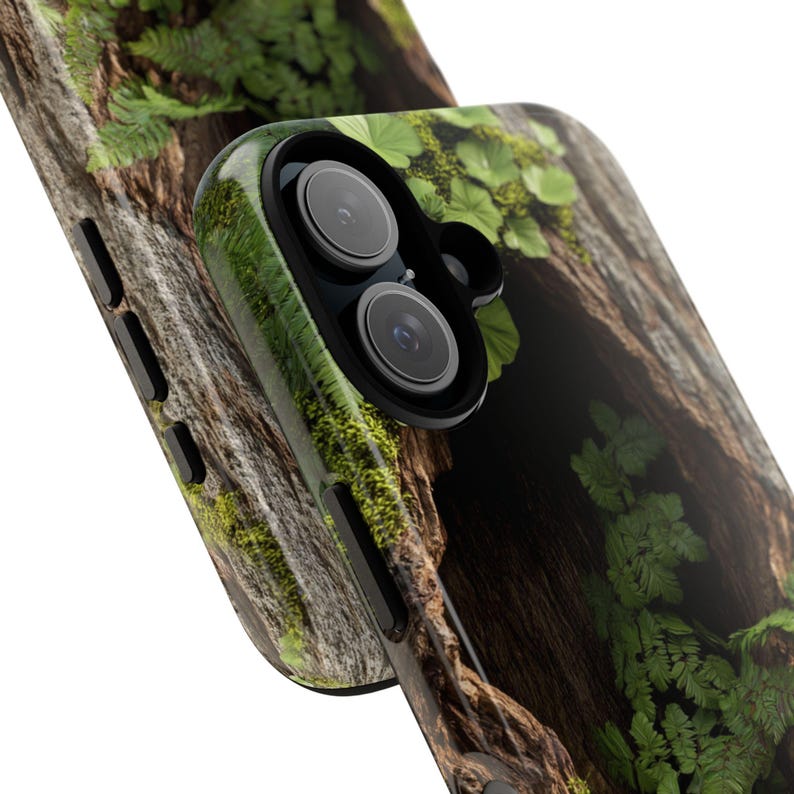 May include: A close-up of a phone case with a forest scene. The case features a dark brown tree trunk with green moss and ferns growing on it. The phone's camera lens is visible in the upper right corner of the image.