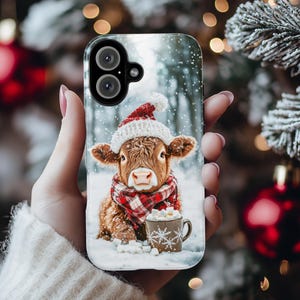 May include: A phone case with a festive design. It features a brown calf wearing a red and white hat and scarf, in a snowy setting. The calf is holding a mug of hot chocolate. The case has a clear border.