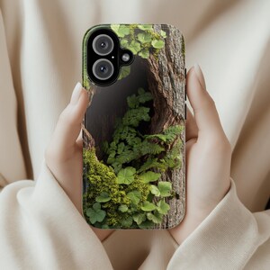 May include: A phone case with a green and brown nature scene. The case features a close-up of a tree trunk with a hole in it, filled with green moss and ferns.