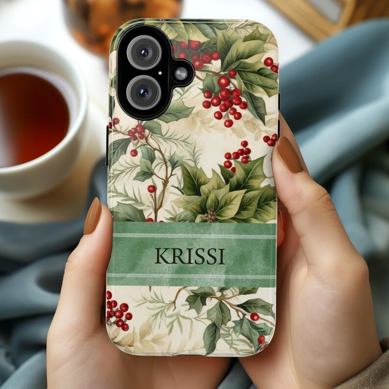 May include: A phone case featuring a festive design with holly leaves and red berries. The case has a green banner with the name "KRISSI" printed on it. The background is a cream color.