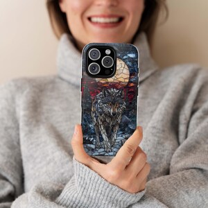 May include: A phone case featuring a stained-glass style design of a wolf under a full moon. The wolf is gray and brown, with a detailed mosaic pattern. The background includes a red and blue forest scene.