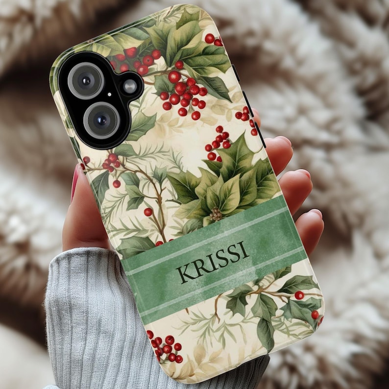 May include: A phone case featuring a festive design with green leaves, red berries, and a cream-colored background. A green banner with the name "KRISSI" is displayed on the case. The case is designed to fit a smartphone.