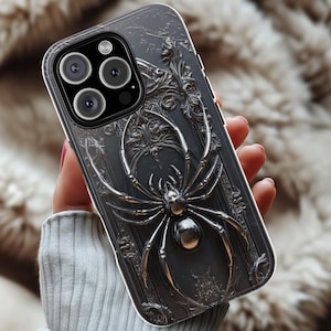 Puede incluir: Carcasa de teléfono negra con un diseño de araña plateada detallada sobre un fondo negro. La carcasa tiene una protuberancia de cámara elevada con tres lentes.