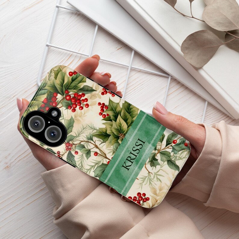 May include: A phone case featuring a botanical design with green leaves, red berries, and the name "KRISSI" in white on a green stripe. The case has a camera cutout and is held in a person's hands.