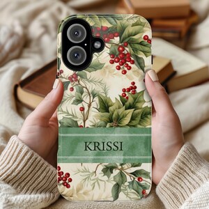 May include: A phone case featuring a festive botanical design with holly leaves and red berries. A green banner at the bottom displays the name "KRISSI" in a bold font. The case has a cream-colored background.