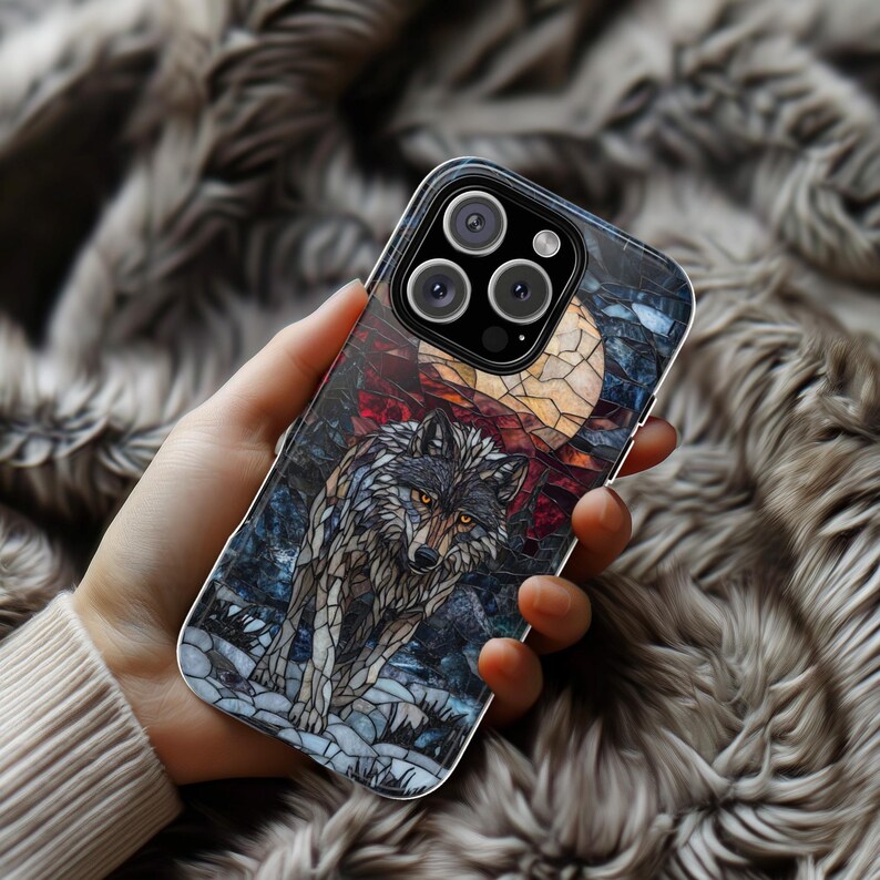 May include: A phone case featuring a wolf design. The artwork depicts a wolf under a full moon, with a stained-glass effect. The color palette includes shades of gray, blue, red, and yellow. The case is held in a hand, with a blurred background.