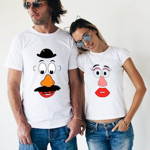 May include: Two white t-shirts with a cartoon face design. One shirt features a Mr. Potato Head face with a black hat, orange nose, and black mustache. The other shirt features a Mrs. Potato Head face with pink lips, eyelashes, and a pink nose.
