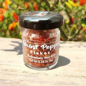 May include: A small glass jar filled with red and orange ghost pepper flakes. The jar has a black lid and a white label that reads "Ghost Pepper Flakes Extremely Hot (!!!) Net Wt 0.28 Oz (8g)"