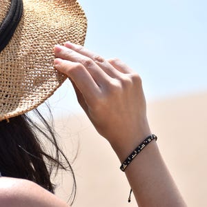Könnte beinhalten: Ein schwarzes Perlenarmband mit goldenen Akzenten wird am Handgelenk getragen. Das Armband besteht aus kleinen, quadratischen Perlen. Eine Person trägt einen Strohhut und hält ihn mit der Hand. Der Hintergrund ist ein Strand mit blauem Himmel.