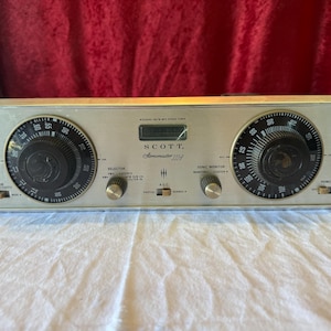 May include: Vintage silver-toned Scott Stereomaster 224A stereo tuner. Features two large dials with black numbers and a central display. Includes knobs for AM, FM, and other functions. The unit is on a white surface.