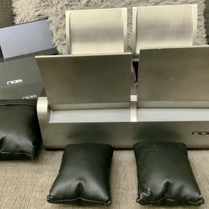 May include: A silver-toned watch display case with two compartments, each with a hinged lid. Includes black leather watch pillows and boxes. The brand name "NOR" is visible on the case.