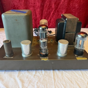 May include: Vintage audio amplifier with a dark gray metal chassis. It features several vacuum tubes, transformers, and cylindrical components. The unit has a rectangular shape with various labels and markings, suggesting it's a classic electronic device.