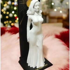 May include: A white statue of a woman in a flowing gown, standing in front of a black rectangular backdrop with a gold sunburst design. The base is black with the inscription "Dalida 1933-1987". The statue is a decorative item.