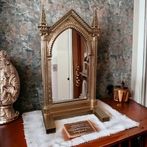 Peut inclure: Un miroir orné de couleur dorée avec un design en arc gothique. Le miroir est posé sur une table en bois avec un tissu blanc et un petit objet doré devant.