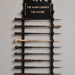 May include: A black wall-mounted display rack holding ten unique wands. The top features a silhouette of a castle and the gold text "THE WAND CHOOSES THE WIZARD." The wands vary in color from light beige to dark brown, featuring intricate carved textures.
