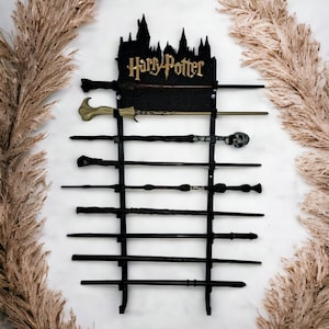 May include: A black wand display rack with the words "Harry Potter" in gold lettering. The rack holds several wands of varying designs. The background is white with decorative dried plants on either side.