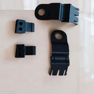 Puede incluir: Se muestran cuatro soportes metálicos negros de diferentes tamaños sobre un fondo neutro. Cada soporte tiene una forma única, que incluye una pieza rectangular con dos agujeros y piezas con un agujero circular y un extremo bifurcado.