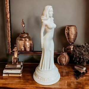 May include: White ceramic statue of a woman in a flowing gown. The statue is on a pedestal with the word "Dali" inscribed on it.