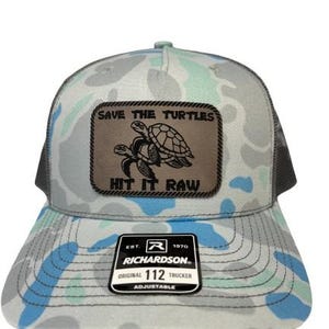Pode incluir: Um chapéu de caminhoneiro de camuflagem cinza e azul com um remendo de couro marrom que diz "Save the Turtles" e "Hit it Raw". O chapéu é ajustável e tem um logotipo Richardson na frente.