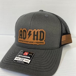 Può includere: Un cappello da camionista grigio e nero con una toppa in pelle marrone che dice "AD HD HIGHWAY TO HEY LOOK, A SQUIRREL". Il cappello ha una parte posteriore in rete nera e una toppa della bandiera americana. Il cappello è etichettato "EST. 1970 RICHARDSON ORIGINAL 112 TRUCKER ADJUSTABLE".
