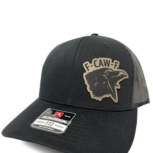 Puede incluir: Gorra de camionero negra Richardson 112 con parte trasera de malla y visera curva. La gorra presenta un parche con un gráfico de cuervo y el texto "F-CAW-F". La gorra es ajustable.