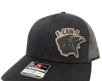 May include: Black Richardson 112 trucker hat with a mesh back and a curved brim. The hat features a patch with a raven graphic and the text "F-CAW-F". The hat is adjustable.