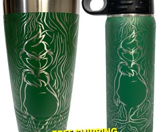 Puede incluir: Un vaso y una botella de agua de acero inoxidable verde con un contorno plateado del Grinch. El Grinch está de pie en un patrón de veta de madera verde y blanco.