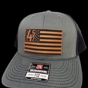 May include: A gray and black trucker hat with a brown leather patch featuring an American flag and the text "457". The hat has a black mesh back and a label that reads "EST. 1970 RICHARDSON ORIGINAL 112 TRUCKER ADJUSTABLE".