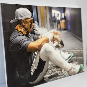 Może przedstawiać: Mężczyzna w szarej czapce z daszkiem z czarno-białym logo siedzi na betonowej podłodze. Ma na sobie czarno-białą koszulkę z napisem "CHAMP" na szyi. Trzyma srebrny puchar i pali papierosa.