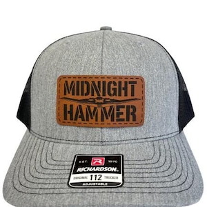 Puede incluir: Gorra de camionero gris con parte trasera de malla negra y visera curva. La gorra presenta un parche de cuero marrón con las palabras "MIDNIGHT HAMMER" en negro. Un pequeño parche en blanco y negro está en la parte delantera.