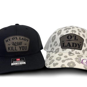 May include: Two trucker caps. One is black with the text "MY O'L LADY WILL KILL YOU" on a leather patch. The other is white with a grey leopard print and the text "O'L LADY" on a leather patch.