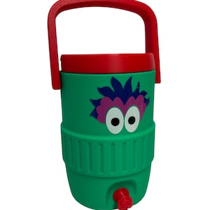 May include: A green beverage cooler with a red handle and lid, featuring a graphic of a purple-haired creature with large white eyes. The cylindrical container has a red spigot at the base for dispensing liquids and a textured middle section.