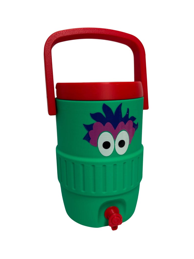 May include: A green beverage cooler with a red handle and lid, featuring a graphic of a purple-haired creature with large white eyes. The cylindrical container has a red spigot at the base for dispensing liquids and a textured middle section.