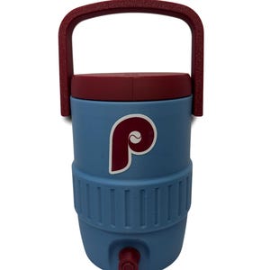 May include: A light blue beverage cooler featuring a maroon lid and a matching maroon handle. The front of the cooler displays a maroon and white letter P logo. A small maroon spigot is located at the bottom center of the cylindrical container.