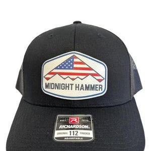 May include: Black trucker hat with a patch featuring an American flag design and the words "MIDNIGHT HAMMER." The hat has a mesh back and an adjustable strap. The Richardson 112 Trucker hat is a classic style.