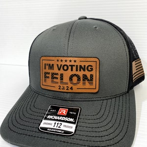 May include: A gray and black trucker hat with a brown leather patch that says "I'M VOTING FELON 2024". The hat is adjustable and has a Richardson 112 label.