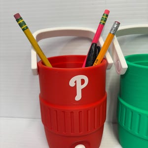 May include: A red cylindrical desk organizer shaped like a miniature beverage cooler with a white handle and a white letter P printed on the front. The container holds two yellow pencils and a black pen, and features a small white spigot at the base.