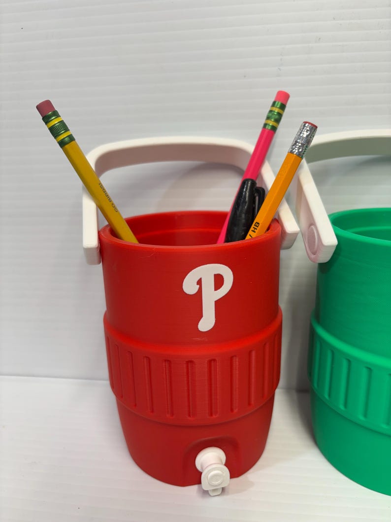 May include: A red cylindrical desk organizer shaped like a miniature beverage cooler with a white handle and a white letter P printed on the front. The container holds two yellow pencils and a black pen, and features a small white spigot at the base.
