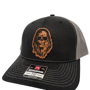 Peut inclure: Casquette de camionneur noire et grise avec un patch en cuir marron d'un personnage avec une barbe et des lunettes de soleil. Le patch indique "112" dessus.