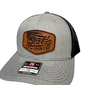 Op de afbeelding: Grijze truckerpet met een zwarte mesh achterkant. De voorkant heeft een bruine leren patch met de tekst "God's Country" en een Amerikaans vlagontwerp. De pet is verstelbaar en heeft een gebogen klep.