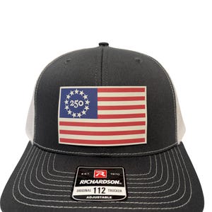 May include: A dark grey trucker cap with a white mesh back and a flat peak. The front displays a patch with a flag design, featuring red and white stripes and a blue field with stars and the number 250. The cap has the Richardson brand label.