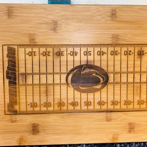 May include: A brown wooden cutting board with a football field design and the words "Penn State" engraved on the side. The board has a handle and a large Nittany Lion logo in the center.