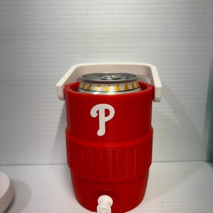 May include: A red cylindrical beverage cooler with a white handle and a white spigot at the base. The front features a white letter P logo. A silver metal beverage can with yellow accents is inserted into the top opening of the cooler.