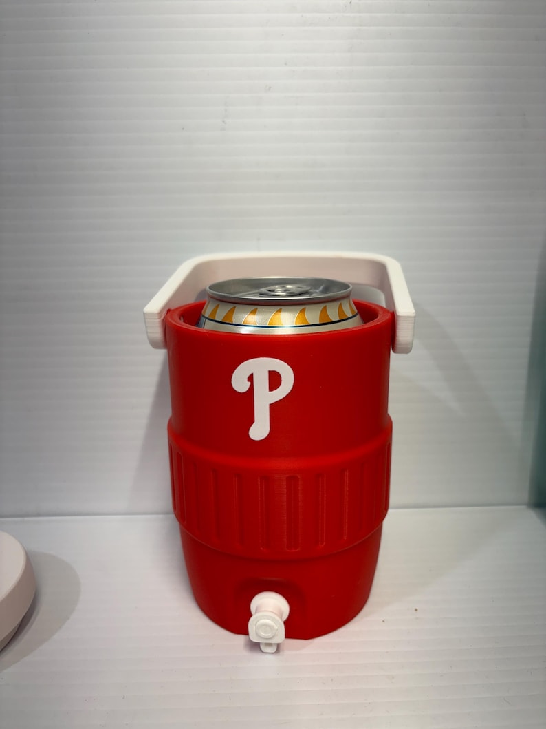 May include: A red cylindrical beverage cooler with a white handle and a white spigot at the base. The front features a white letter P logo. A silver metal beverage can with yellow accents is inserted into the top opening of the cooler.