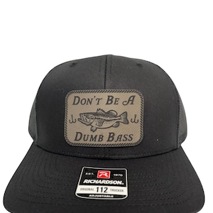 Op de afbeelding: Zwarte truckerpet met een mesh achterkant en een gebogen klep. De patch aan de voorkant leest "DON'T BE A DUMB BASS" met een visafbeelding. De pet is verstelbaar en heeft het Richardson-merklogo.