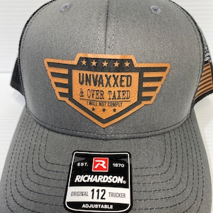 Puede incluir: Un gorro de camionero gris y negro con un parche de cuero marrón que dice "UNVAXXED & OVER TAXED I WILL NOT COMPLY". El gorro es ajustable y tiene una etiqueta Richardson 112.