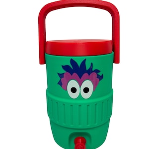 May include: A green beverage cooler with a red lid and a red carrying handle. The front features a graphic of a purple-haired character with large white eyes. A red spigot is located at the bottom of the cylindrical container for dispensing liquid.
