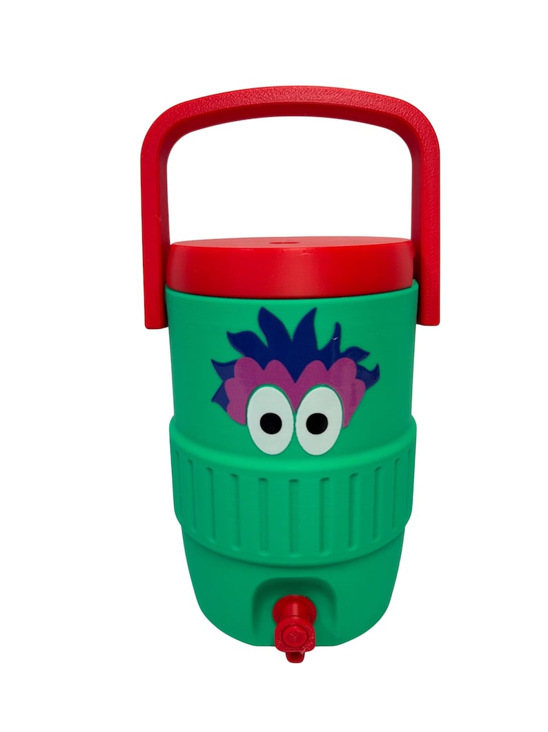 May include: A green beverage cooler with a red lid and a red carrying handle. The front features a graphic of a purple-haired character with large white eyes. A red spigot is located at the bottom of the cylindrical container for dispensing liquid.