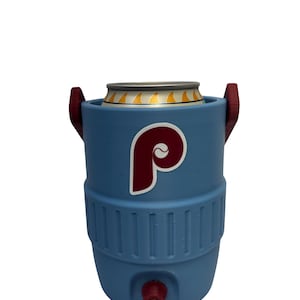 May include: A light blue beverage cooler shaped like a miniature water jug, featuring a maroon and white letter P logo on the front. The cooler has two maroon side handles, a maroon spigot at the base, and holds a silver aluminum beverage can.